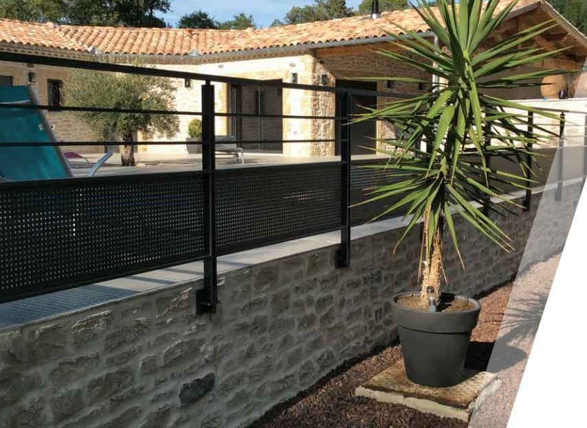 Terrasse en pierre avec balustrade noire, palmier en pot et maison au toit de tuiles sous un ciel bleu
