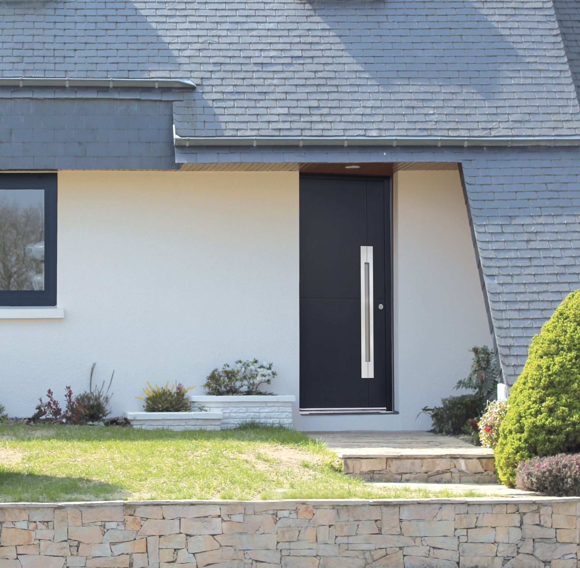 Entrée d'une maison moderne avec porte sombre, mur blanc, toit en ardoise et petite pelouse à l'avant.