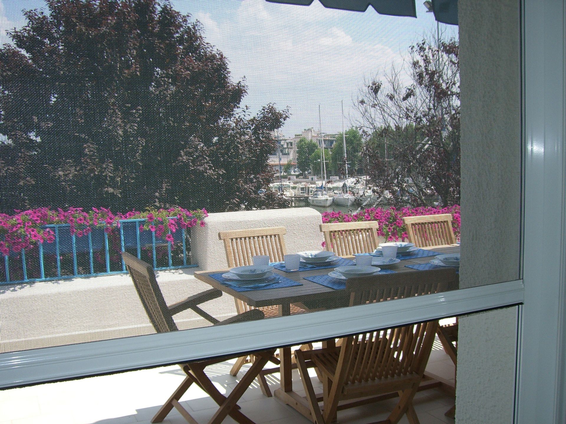 Table et chaises de patio vues par une fenêtre, avec des fleurs et un jardin au-delà.