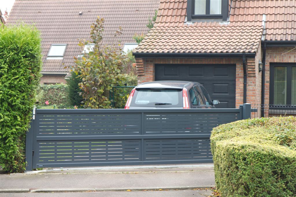 Maison avec une voiture sombre derrière un portail gris et des haies taillées dans la cour avant