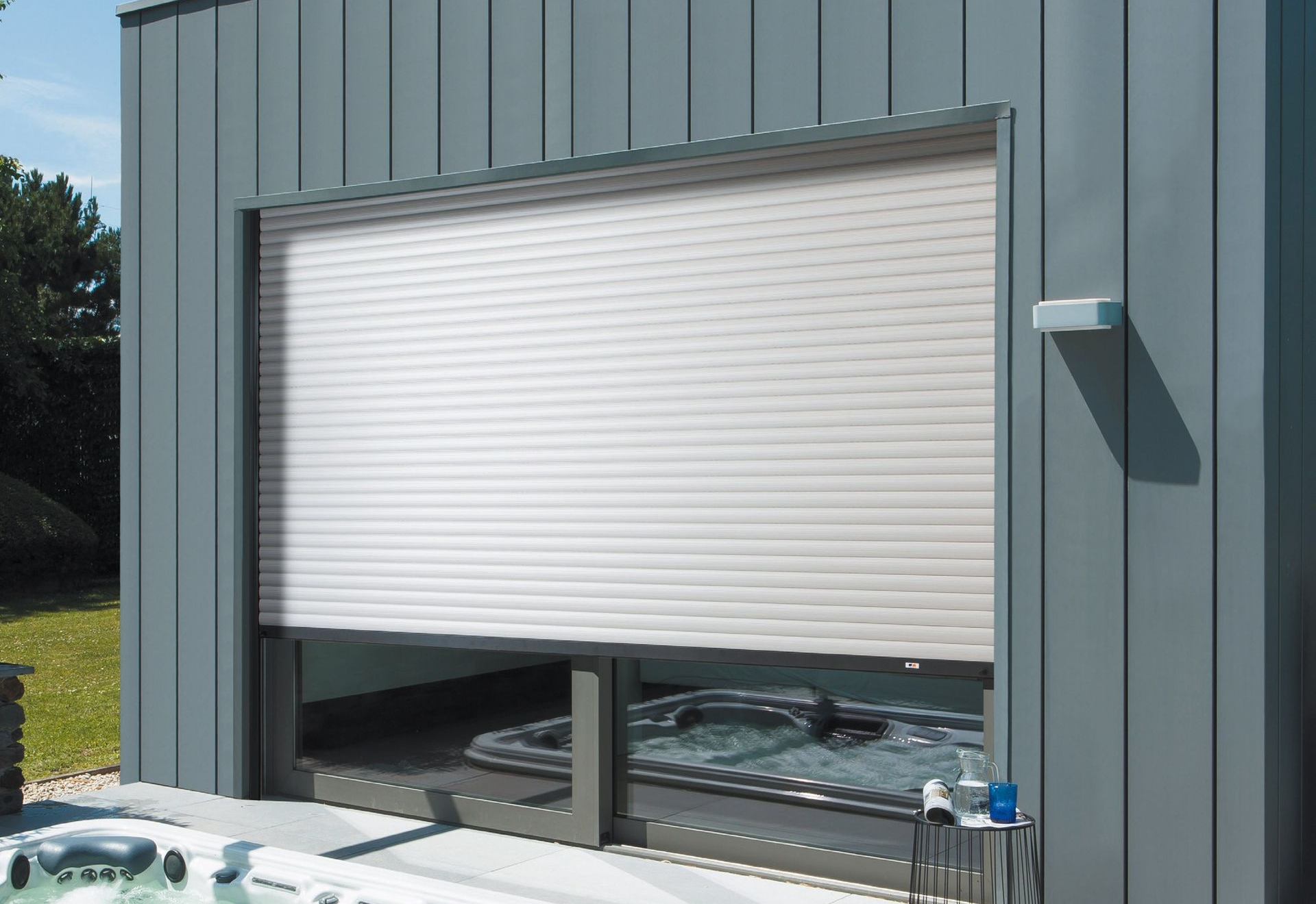 Porte sectionnelle blanche sur un bâtiment métallique gris, partiellement ouverte, laissant apparaître des équipements extérieurs en dessous.