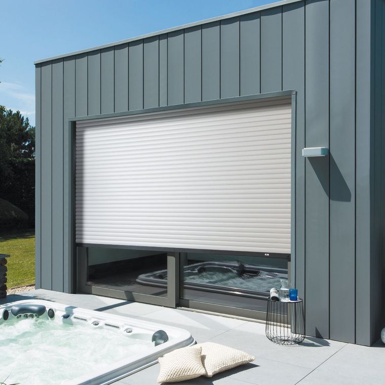 Porte sectionnelle blanche fermée sur un bâtiment gris, à côté d'un jacuzzi et d'une terrasse.