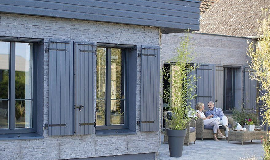Patio moderne avec volets gris, plantes en pot et deux personnes assises à une table extérieure