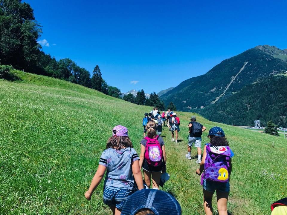 Un gruppo di escursionisti cammina in salita su un sentiero erboso ai piedi di una montagna, sotto un cielo azzurro brillante.