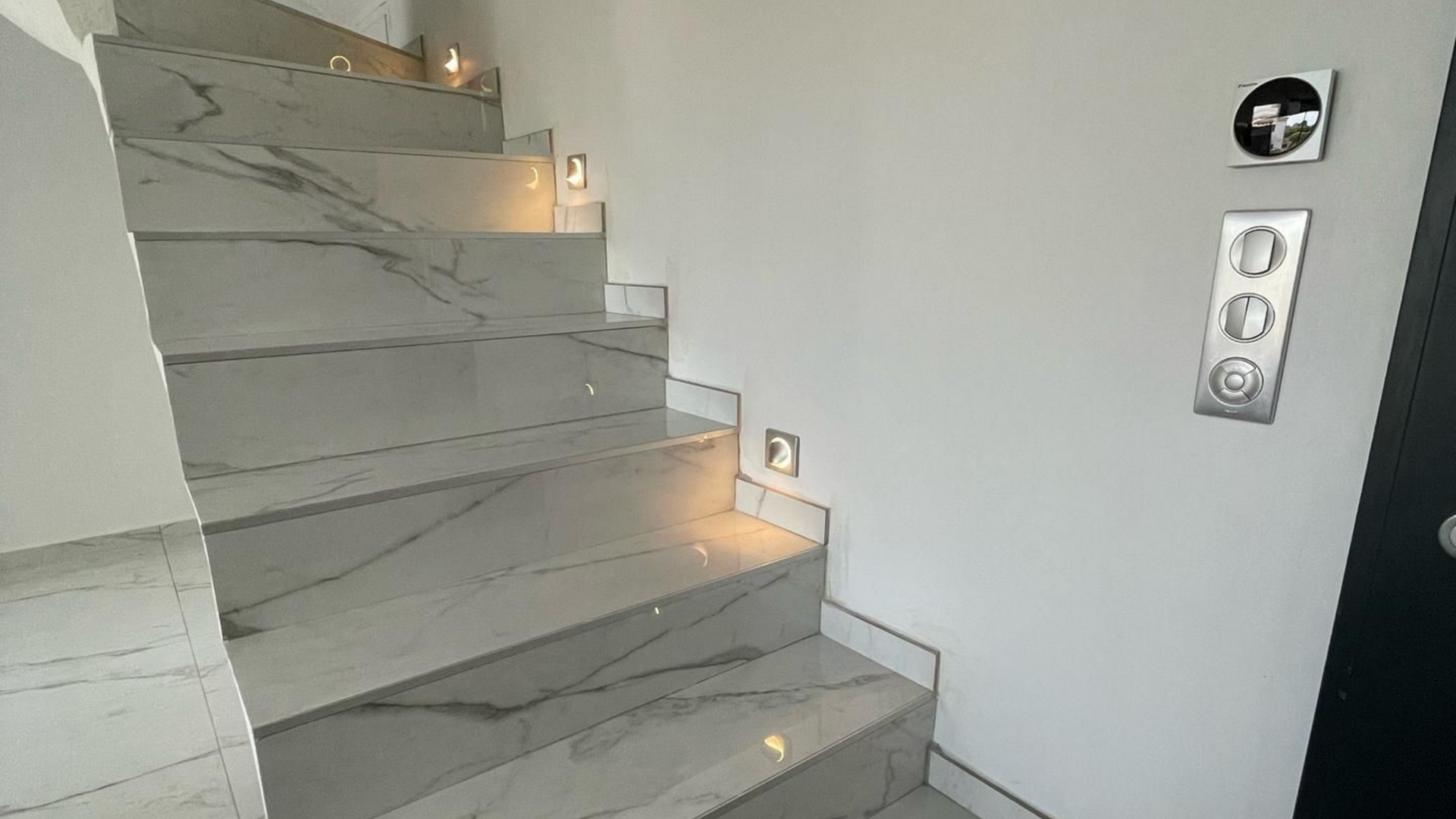 Escalier avec marches imitation marbre, mur avec luminaires, tableau électrique et porte noire.