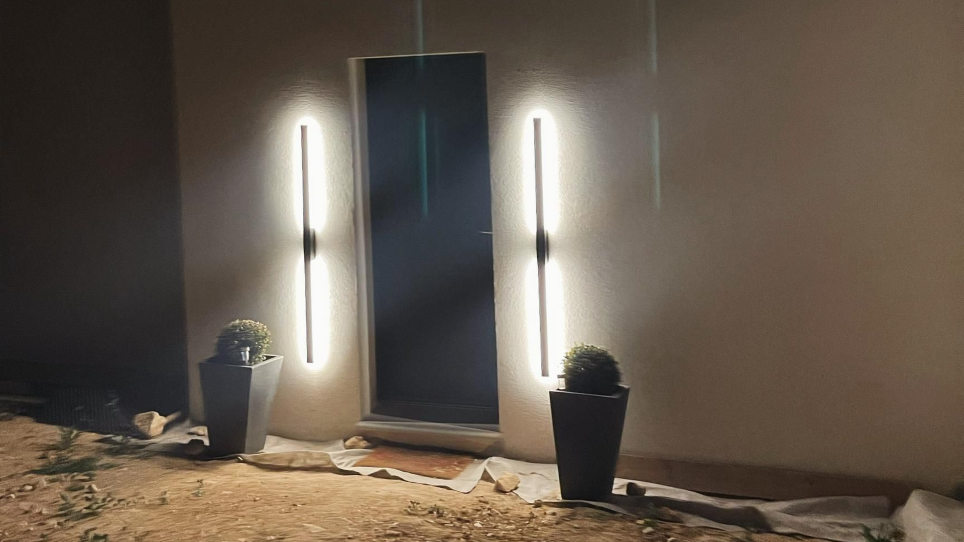 Deux lampes verticales illuminées encadrent une porte sombre, avec des plantes en pot en dessous. Ambiance du soir.