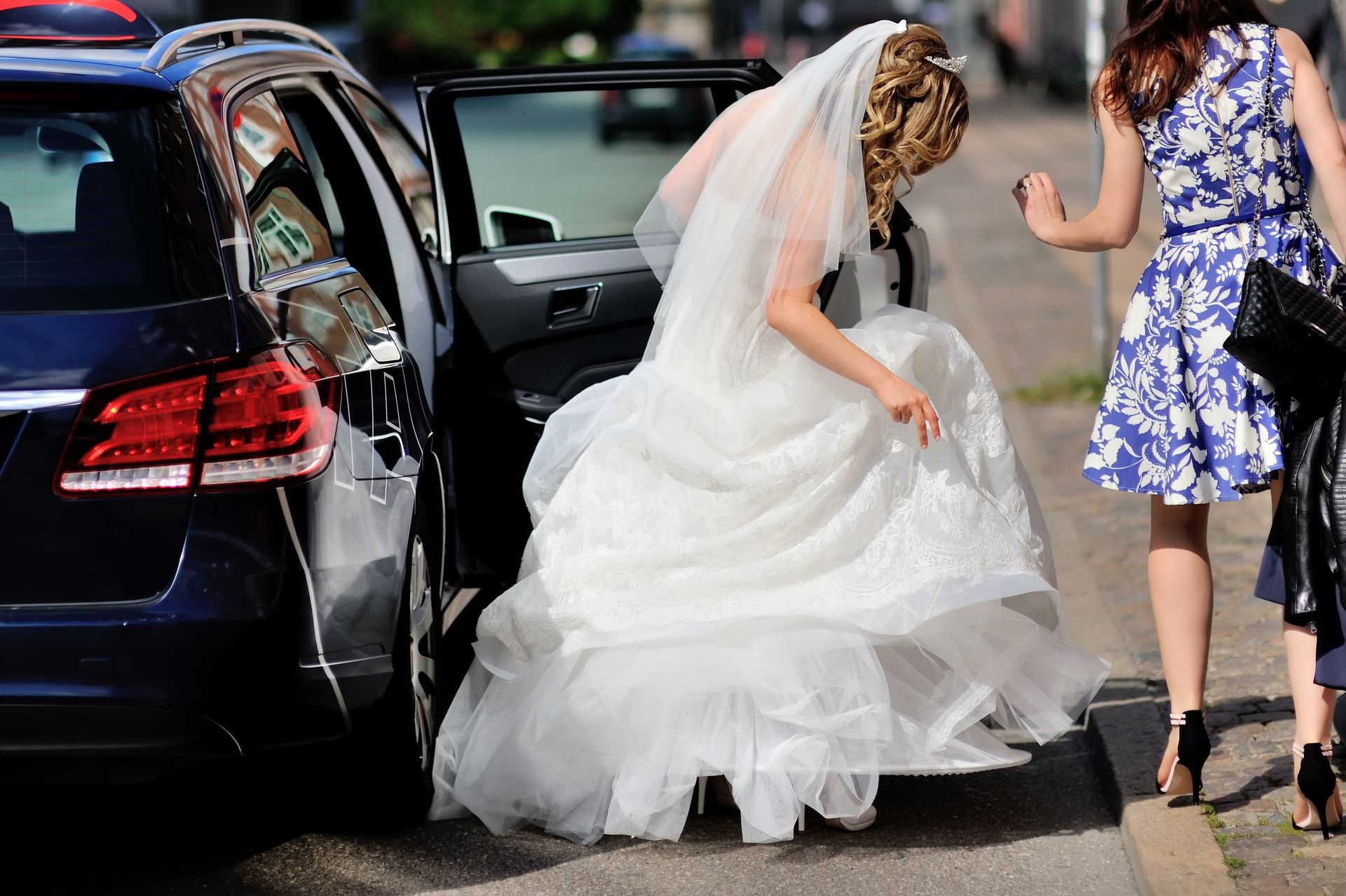Une mariée en robe blanche descend d'une voiture noire, une femme en robe bleue à proximité.