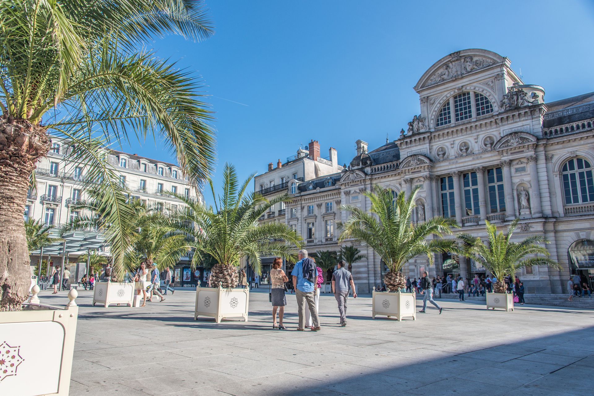 Place ensoleillée avec palmiers et bâtiment historique orné sous un ciel bleu, avec des passants à proximité