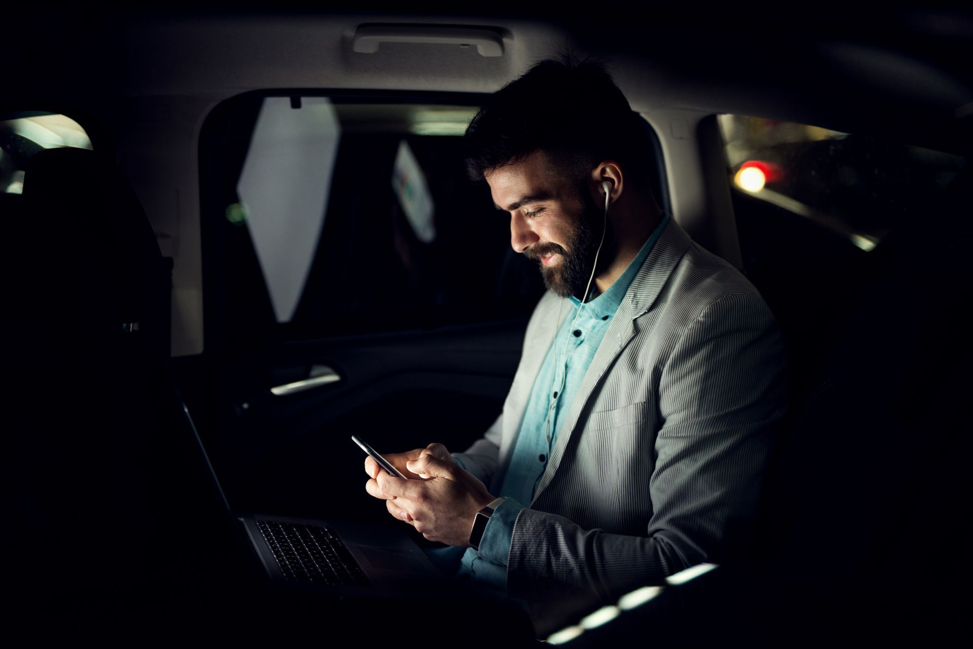 Un homme assis dans une voiture la nuit, regardant un smartphone avec un ordinateur portable ouvert à côté de lui.