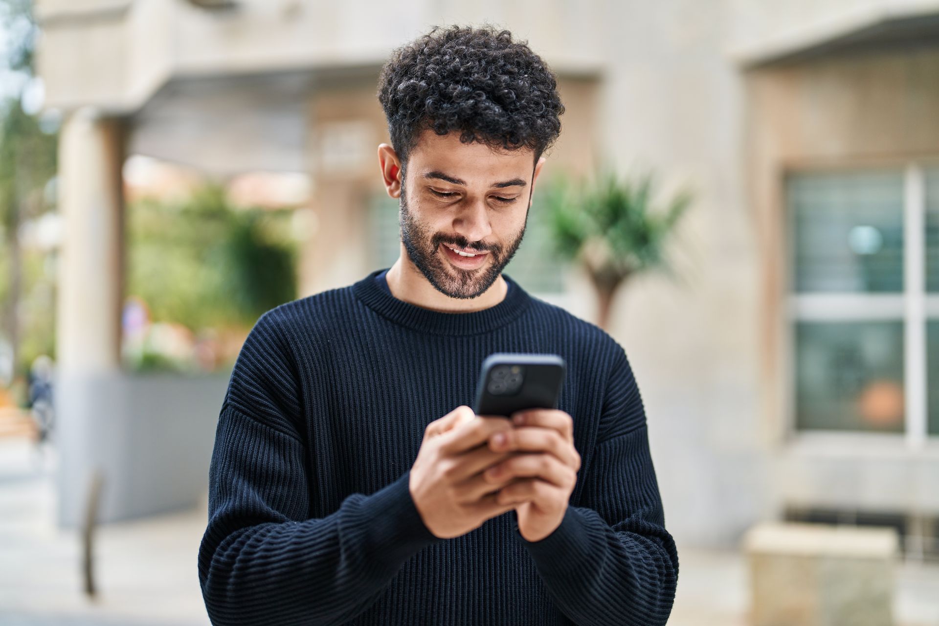 Personne en pull foncé souriant en regardant un smartphone à l'extérieur.