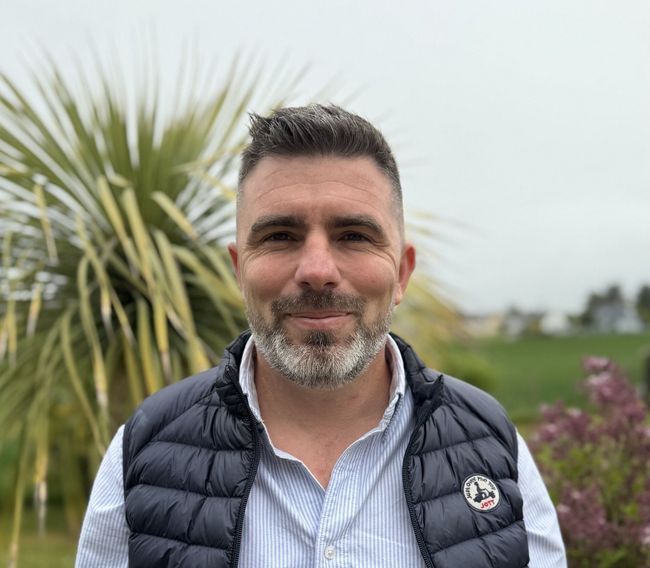 Un homme souriant, vêtu d'un gilet bleu marine et d'une chemise à rayures, se trouve à l'extérieur, avec des plantes et des fleurs en arrière-plan.