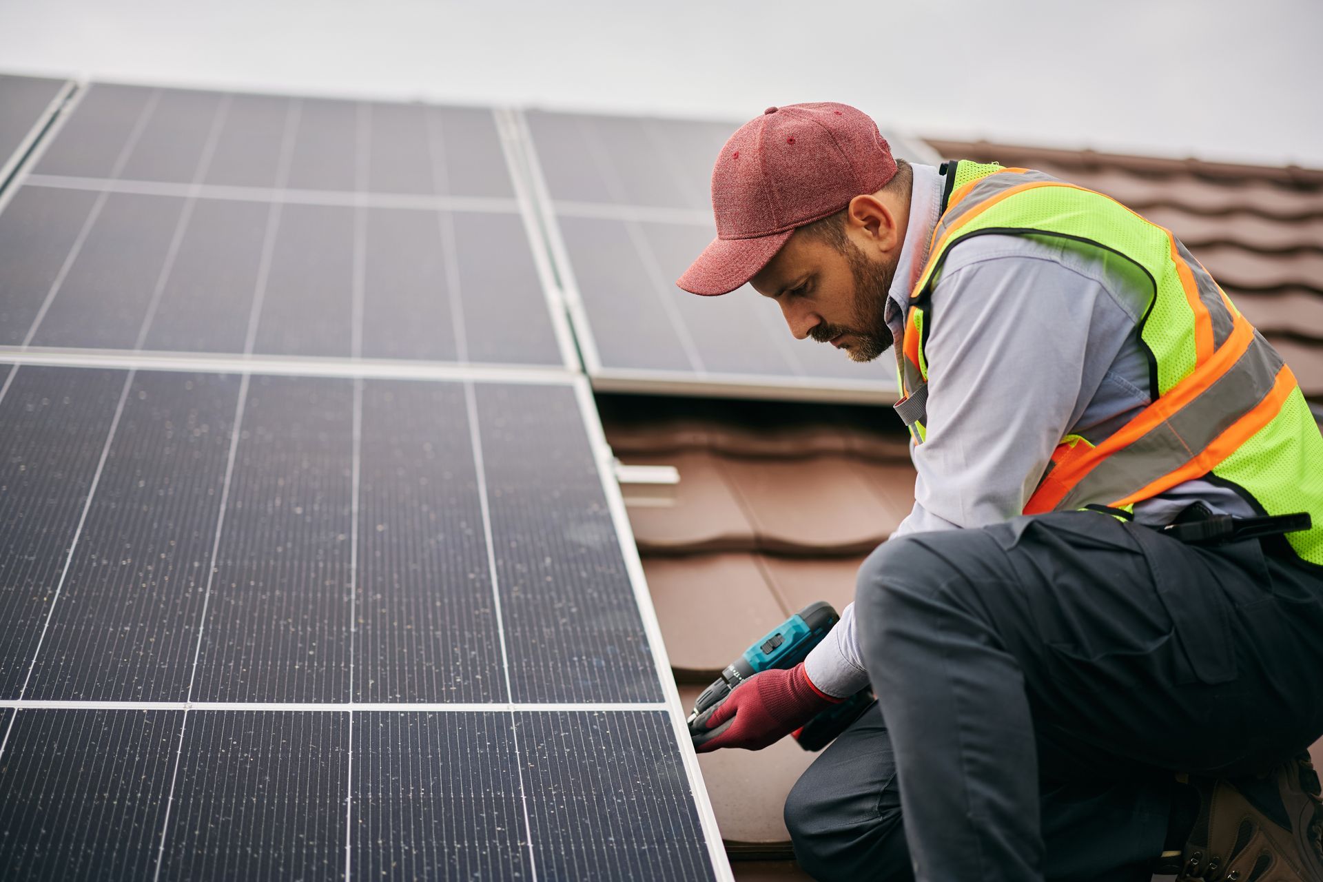 Un technicien portant un gilet haute visibilité et une casquette rouge utilise une perceuse pour installer des panneaux solaires sur un toit de tuiles.