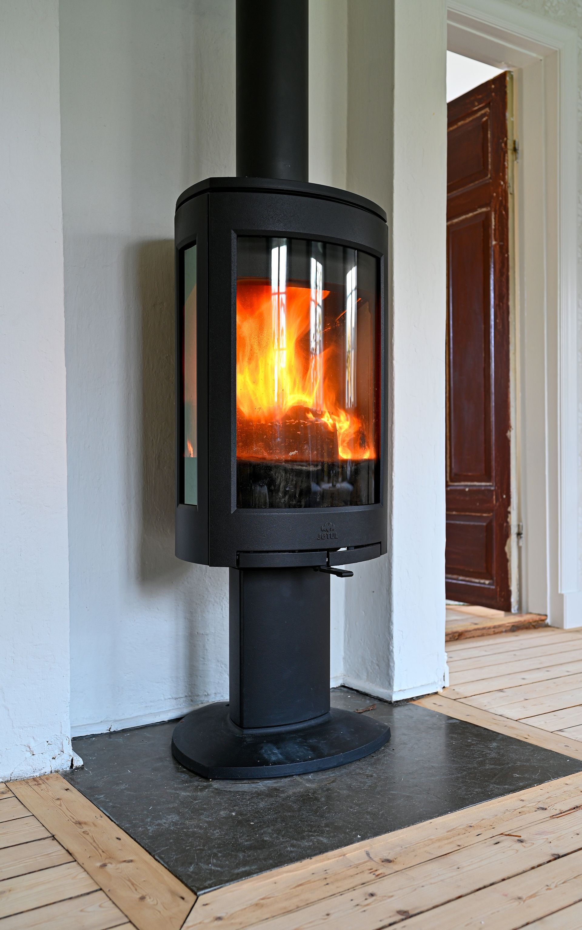 Un poêle à bois cylindrique noir, à porte vitrée et aux flammes vives, posé sur un foyer sombre dans une pièce.