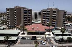 Dos edificios marrones de varios pisos flanquean una plaza central con tiendas. Se ven palmeras y coches aparcados.