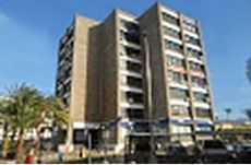 Edificio beige de varios pisos con tiendas en la planta baja, palmeras al frente y cielo azul.