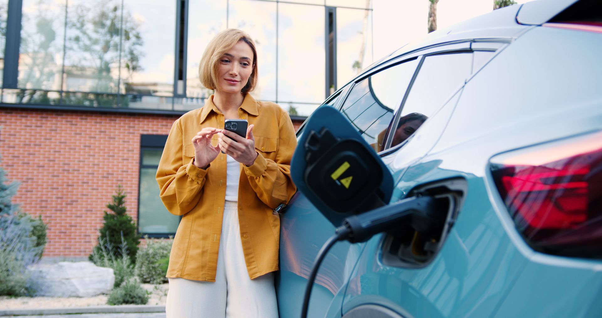 Une personne recharge une voiture électrique bleue tout en utilisant son téléphone à l'extérieur, près d'un bâtiment en briques.