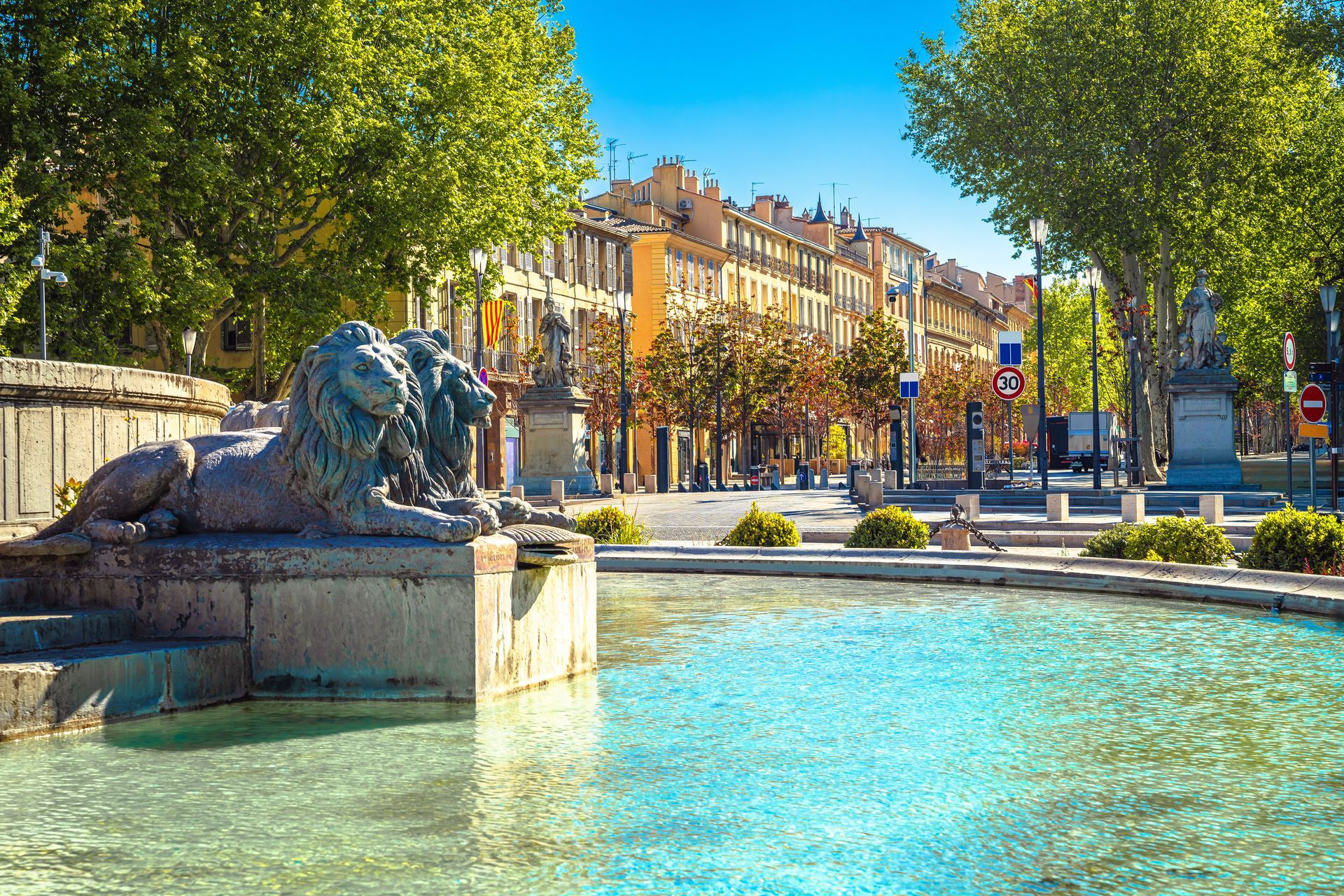 Fontaine urbaine ensoleillée aux eaux turquoise, sculpture de lion en pierre et place bordée d'arbres et de bâtiments historiques