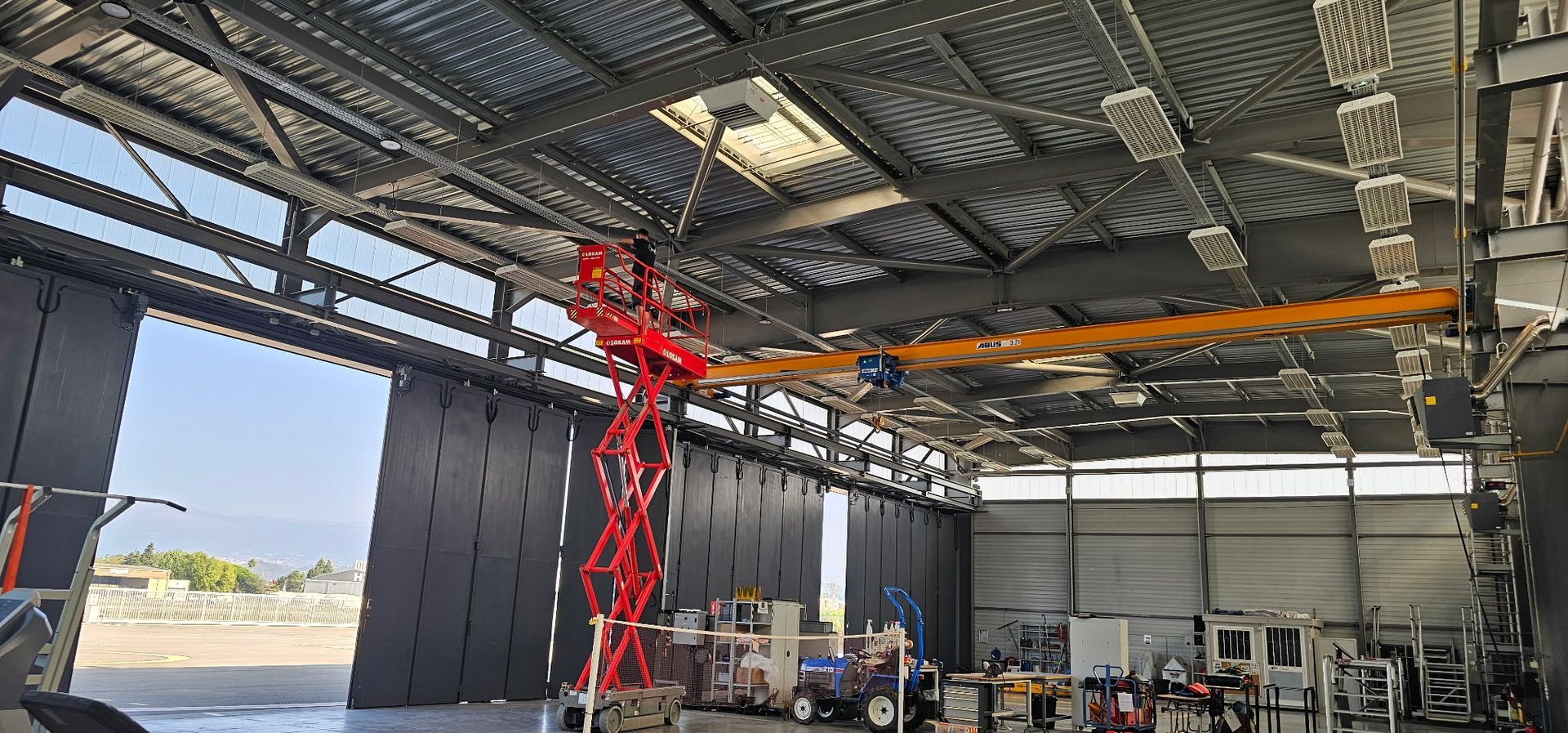 Chantier de construction intérieur avec un portique orange et une nacelle élévatrice rouge sous un toit métallique