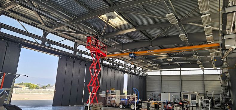 Grand atelier industriel intérieur avec une nacelle élévatrice rouge et un pont roulant orange sous un toit en tôle ondulée