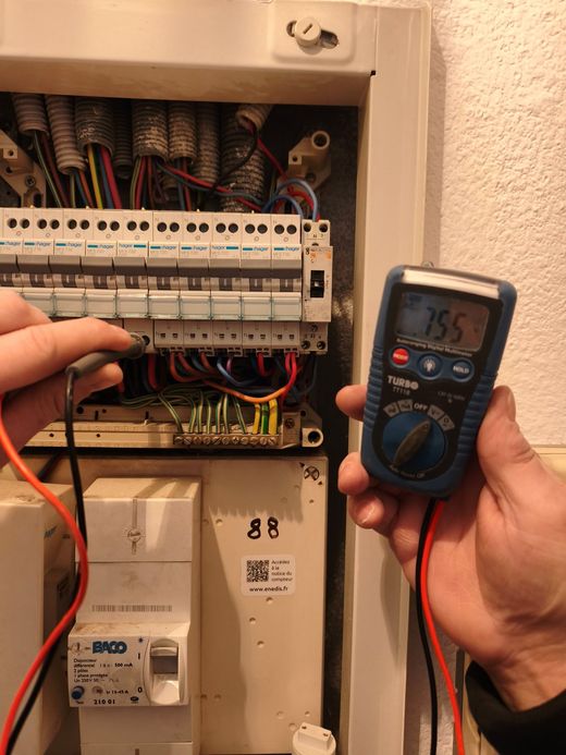 Des mains testent un tableau électrique à l'aide d'un multimètre et de sondes.