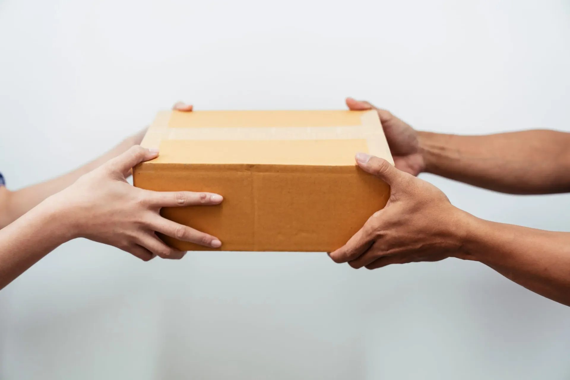 Dos manos pasando una pequeña caja de cartón marrón sobre un fondo blanco.