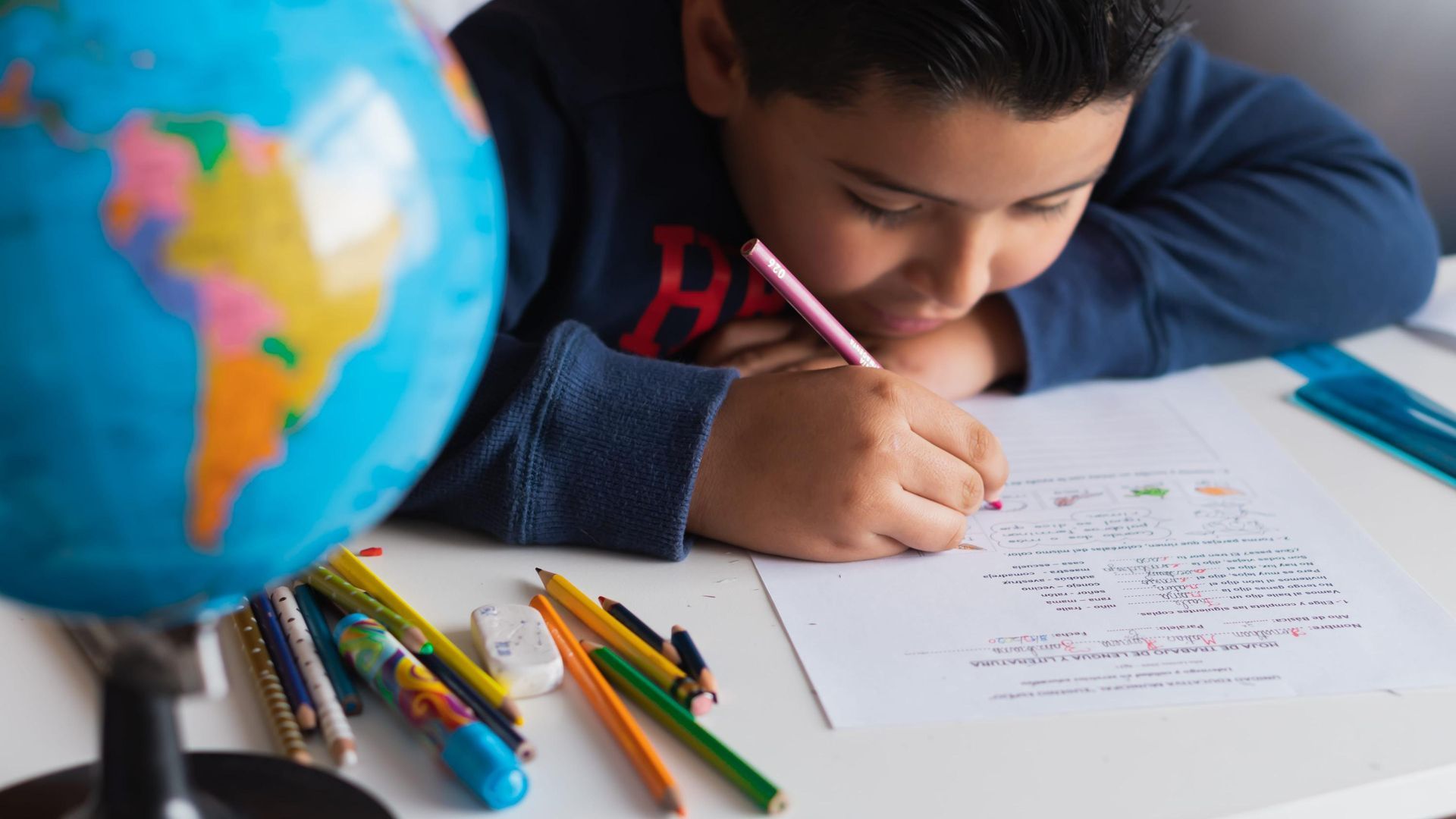Una niña escribe con un lápiz rosa en un escritorio, con lápices de colores y un globo terráqueo en primer plano.