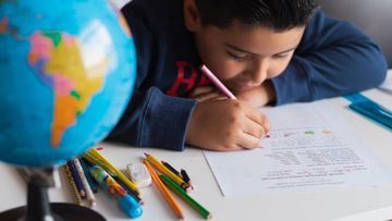 Una niña escribe con un lápiz rosa en un escritorio, con lápices de colores y un globo terráqueo en primer plano.