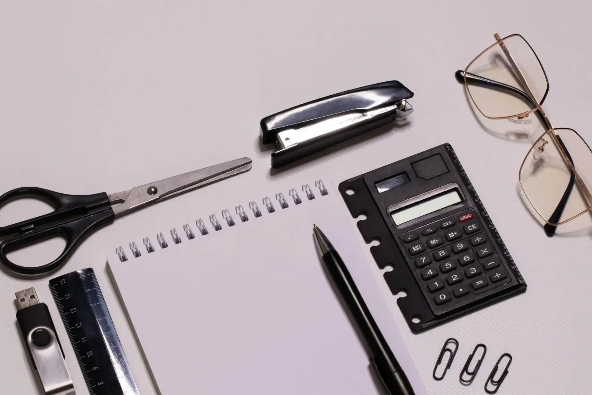 Material de oficina sobre un escritorio claro: tijeras, grapadora, cuaderno, calculadora, bolígrafo, clips y gafas.