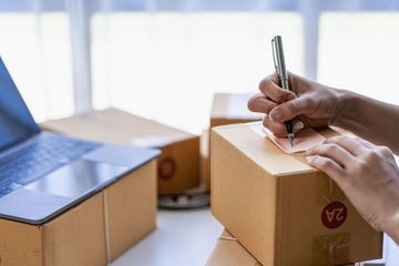 Manos etiquetando una caja de cartón para envíos junto a un ordenador portátil abierto sobre un escritorio.