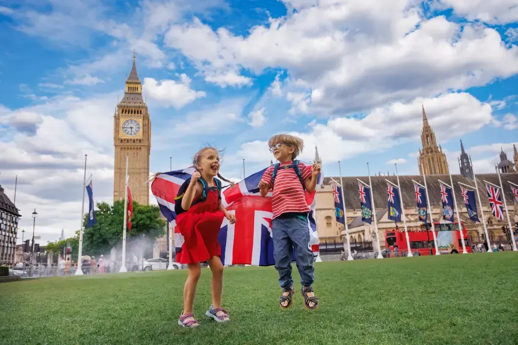 Dos niños vestidos de rojo y azul están de pie sobre un césped, con el Big Ben y el Parlamento al fondo.