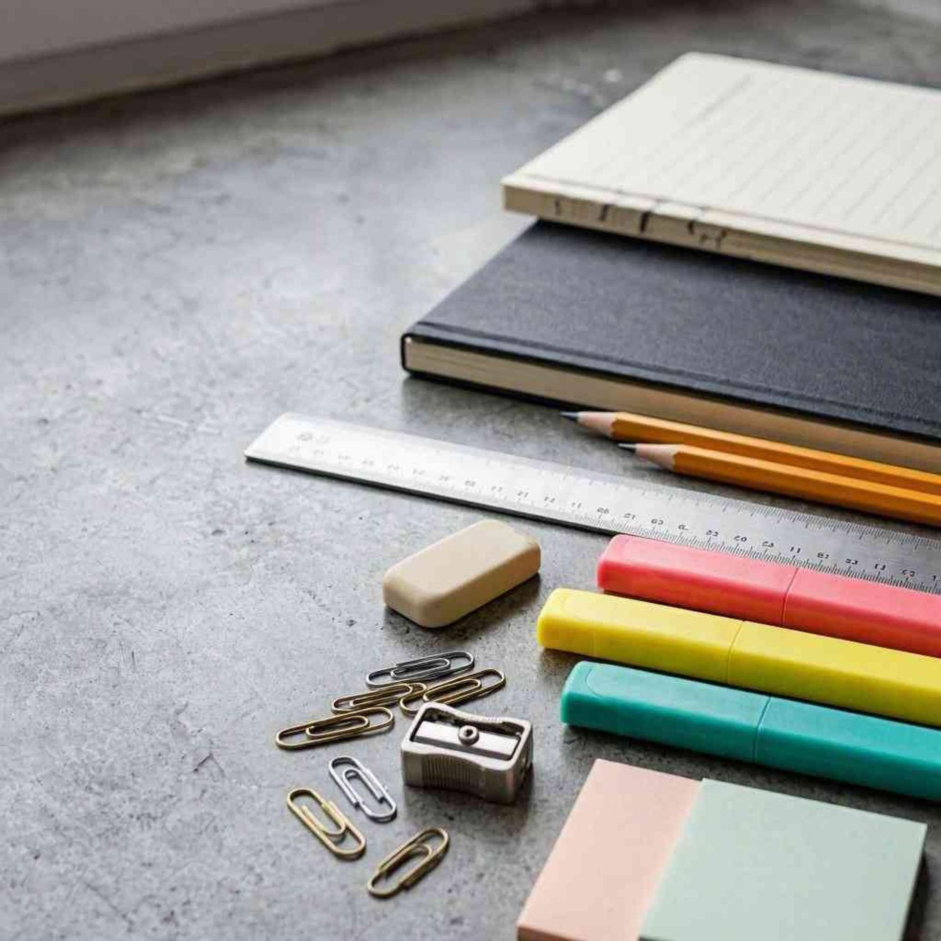 Material de oficina y cuadernos dispuestos sobre una mesa gris, con bolígrafos de colores, clips y una regla.