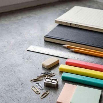 Material de oficina y cuadernos dispuestos sobre una mesa gris, con bolígrafos de colores, clips y una regla.