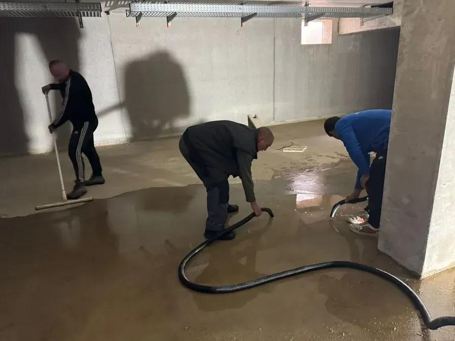 Drei Personen reinigen mit einem Schlauch und einem Abzieher einen nassen Kellerboden aus Beton