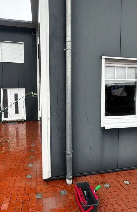 Graue Gebäudefassade mit weiß eingefasstem Fenster, Fallrohr und roter Backsteinterrasse mit einem umgestürzten Pflanzgefäß.