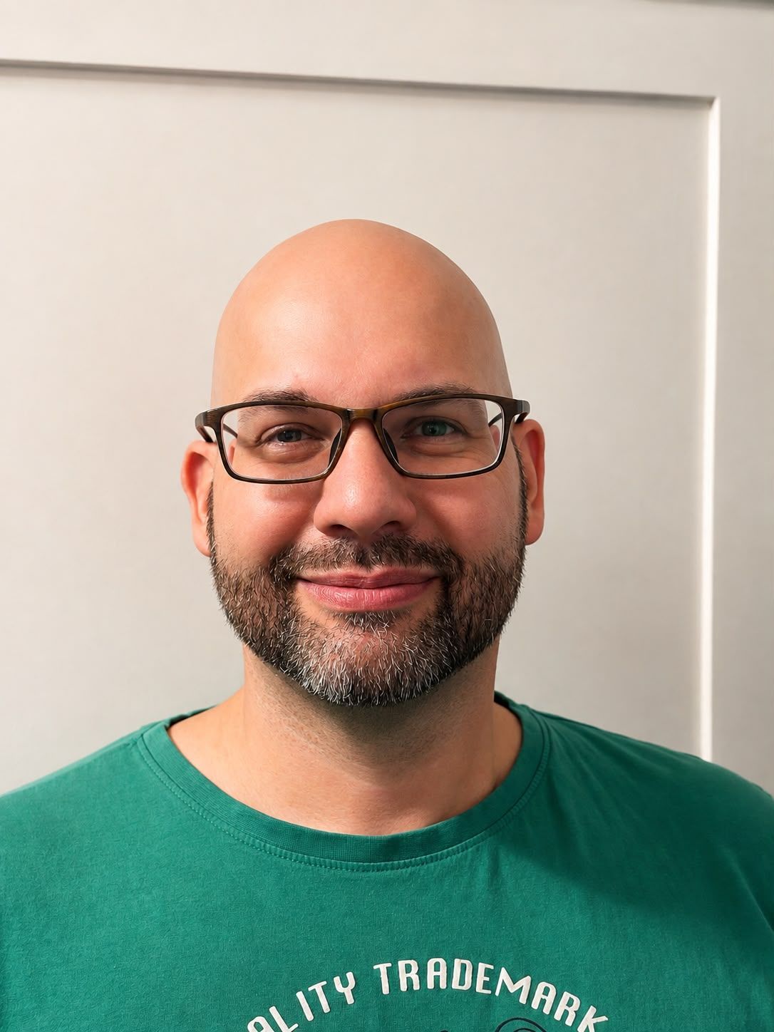 Lächelnder glatzköpfiger Mann mit Brille in einem grünen T-Shirt vor einer weißen Wand
