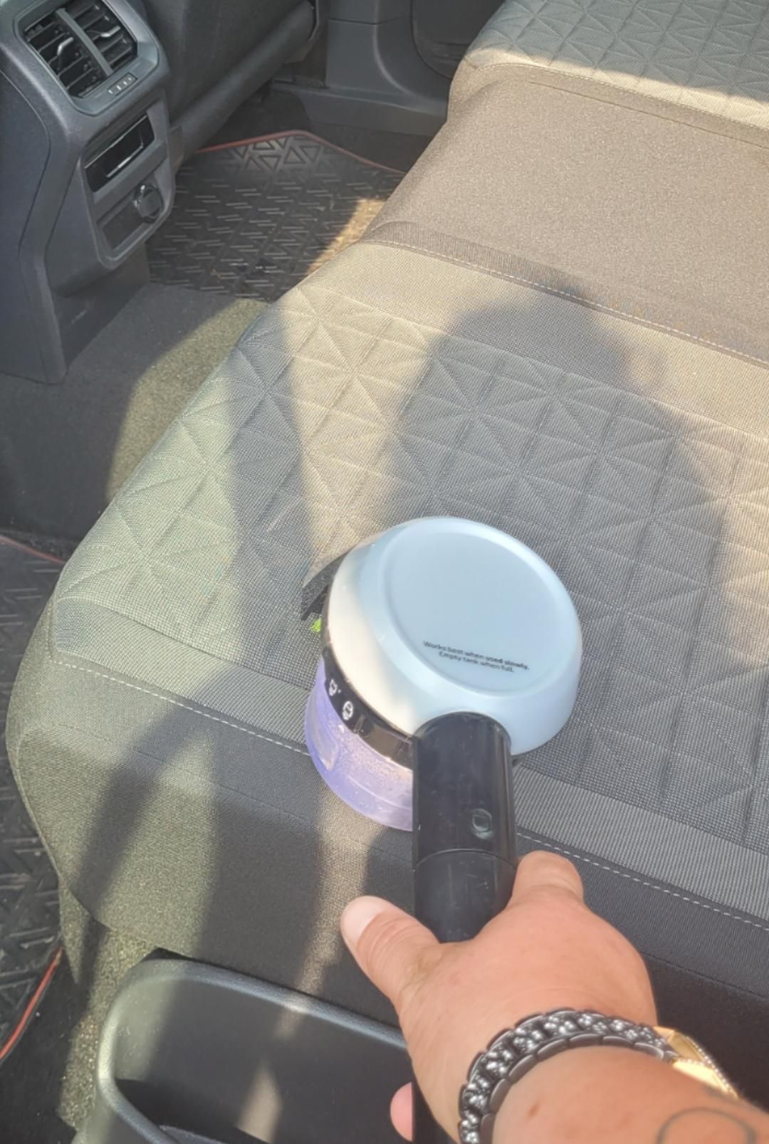 Eine Hand hält einen kleinen Akku-Staubsauger und reinigt damit den Stoffbezug des Vordersitzes im Auto