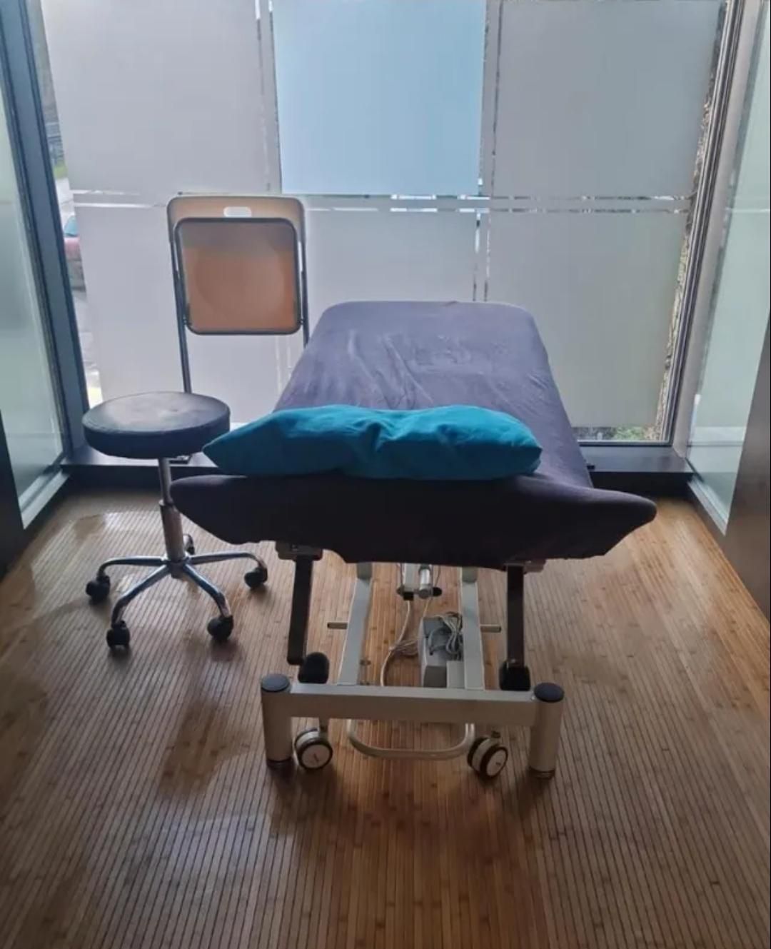 Massageliege mit türkisfarbenem Kopfkissen in einem hellen Raum, neben einem Rollhocker und einem Stuhl.