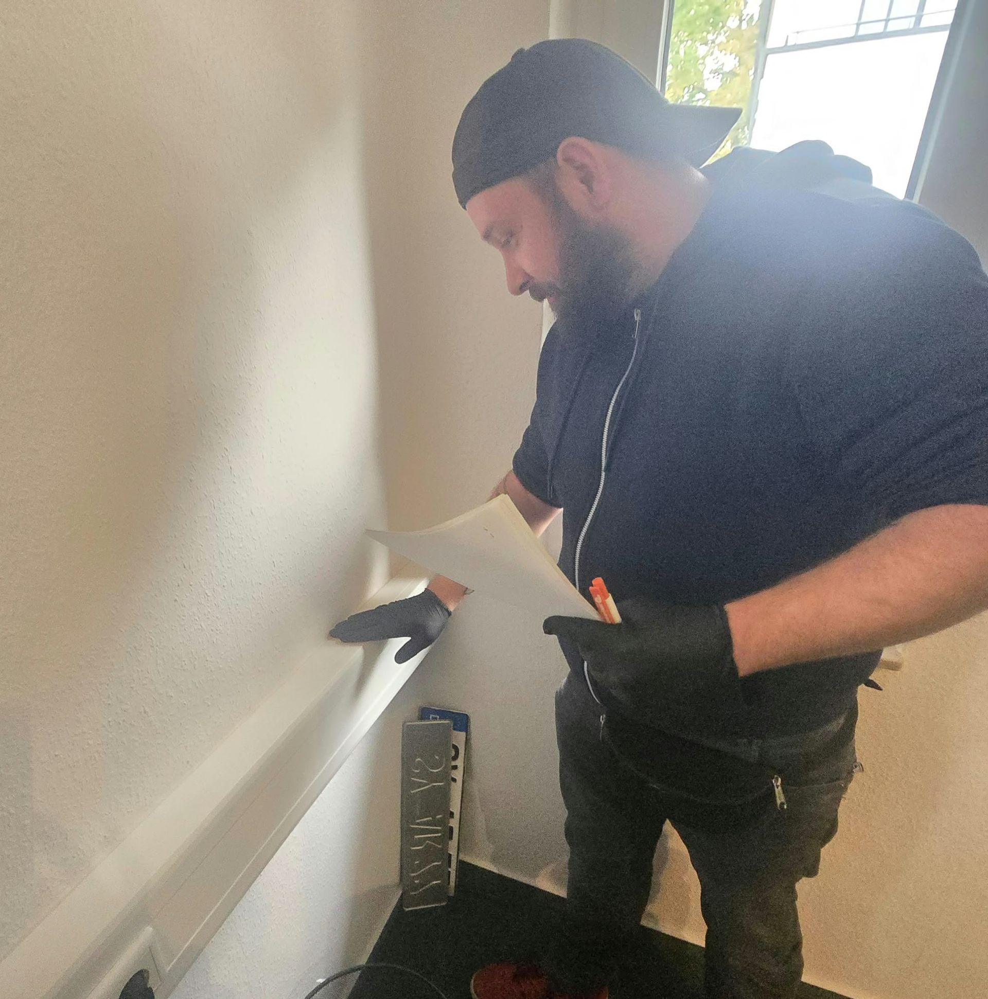Ein Arbeiter untersucht mit einem Werkzeug und einem Blatt Papier in der Hand eine Wand neben einem Treppenhaus.