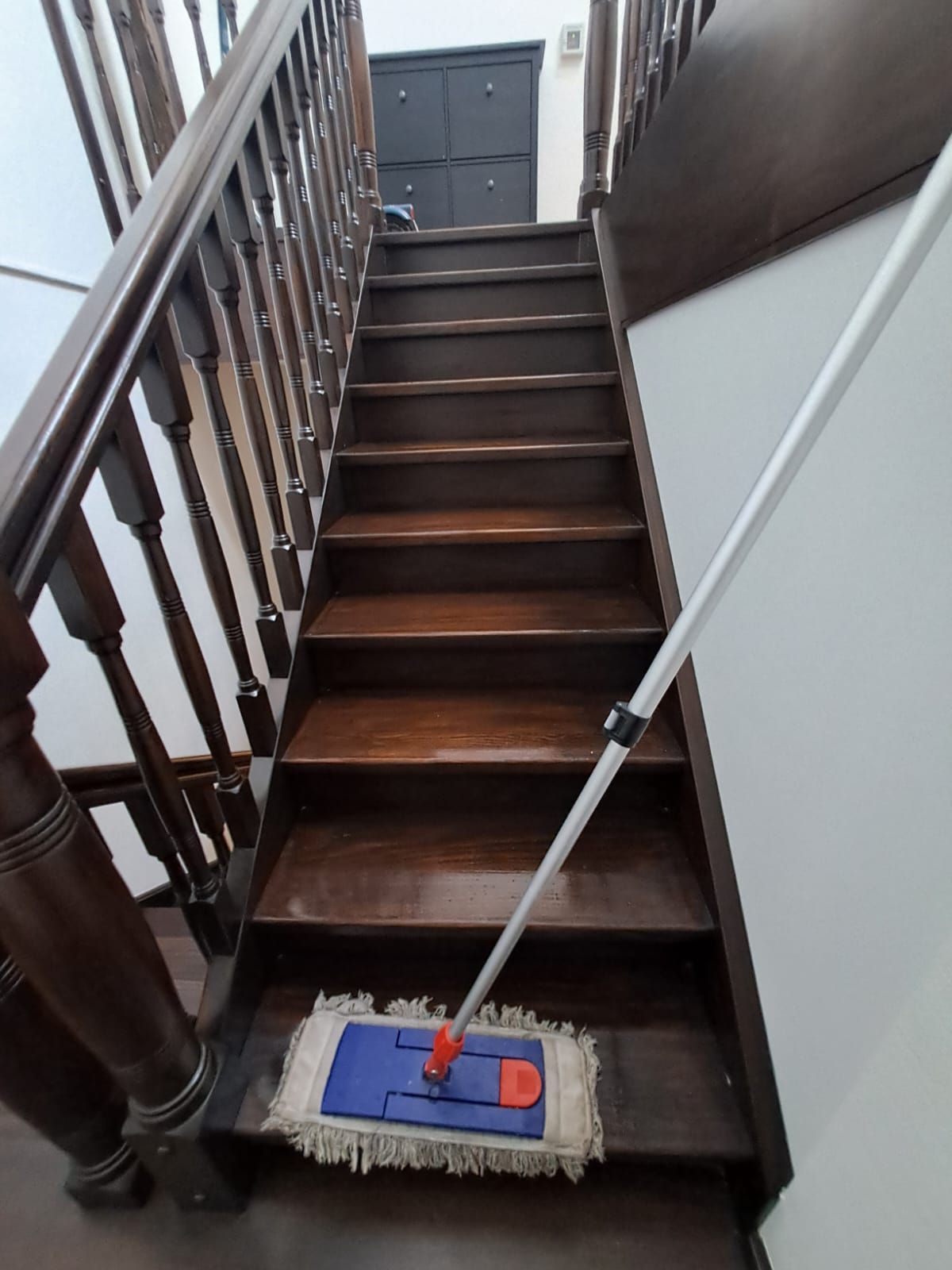 Eine dunkle Holztreppe mit einem Wischmopp und Eimer reinigen; daneben eine weiße Wand und ein Treppengeländer