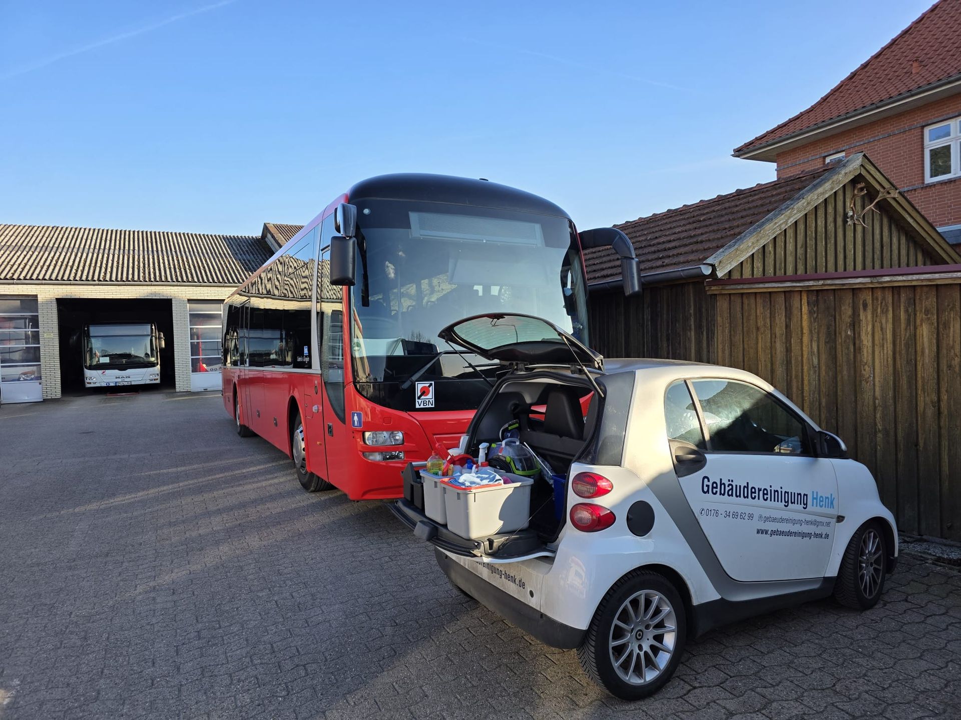 Ein silberner Smart parkt vor einem roten Reisebus. Beide stehen auf einem Parkplatz neben Wohnhäusern unter klarem Himmel