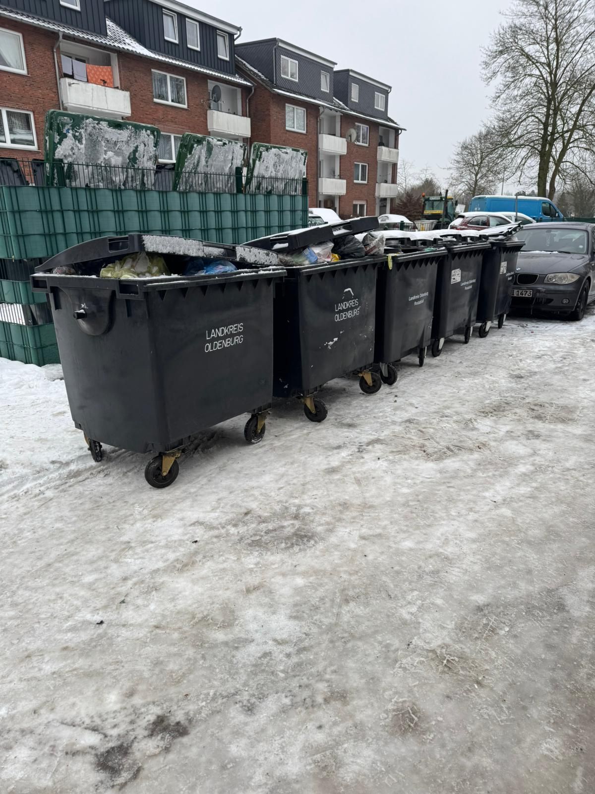 Reihe schwarzer Mülltonnen auf einem schneebedeckten Gehweg neben Wohnhäusern und geparkten Autos