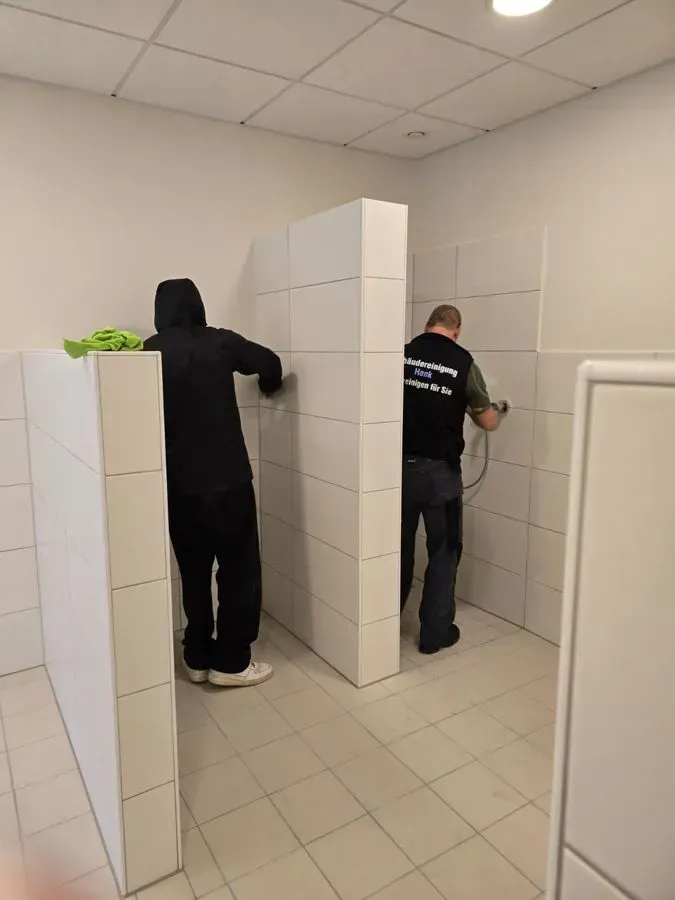 Zwei Personen in einer Toilettenkabine mit weißen Fliesen.