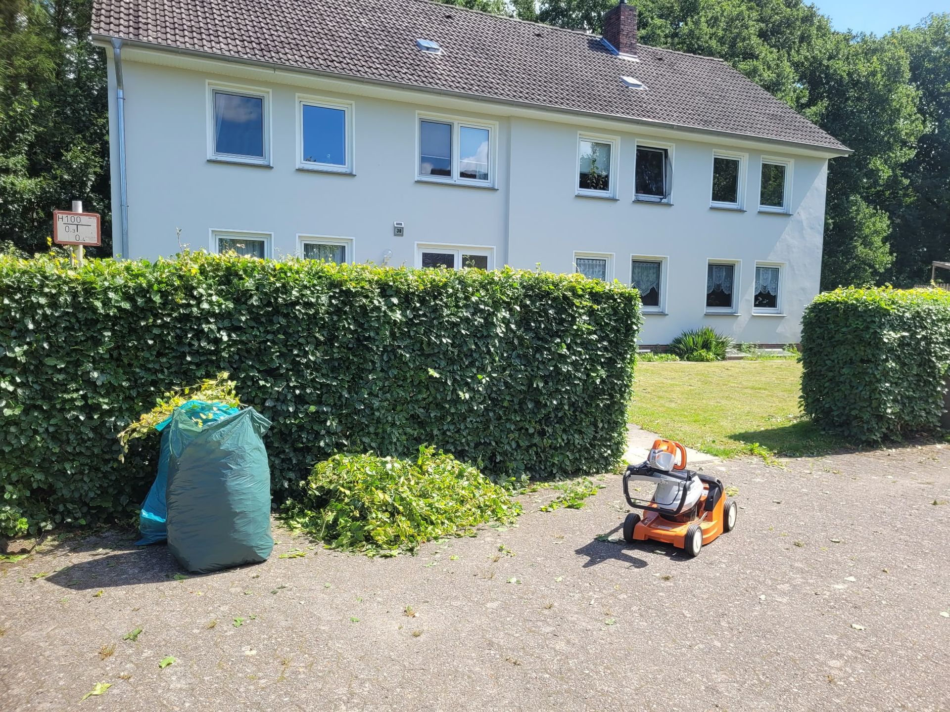 Haus und Hecken mit einer Schubkarre und einem Gartengerät auf einer Schotterauffahrt unter sonnigem Himmel