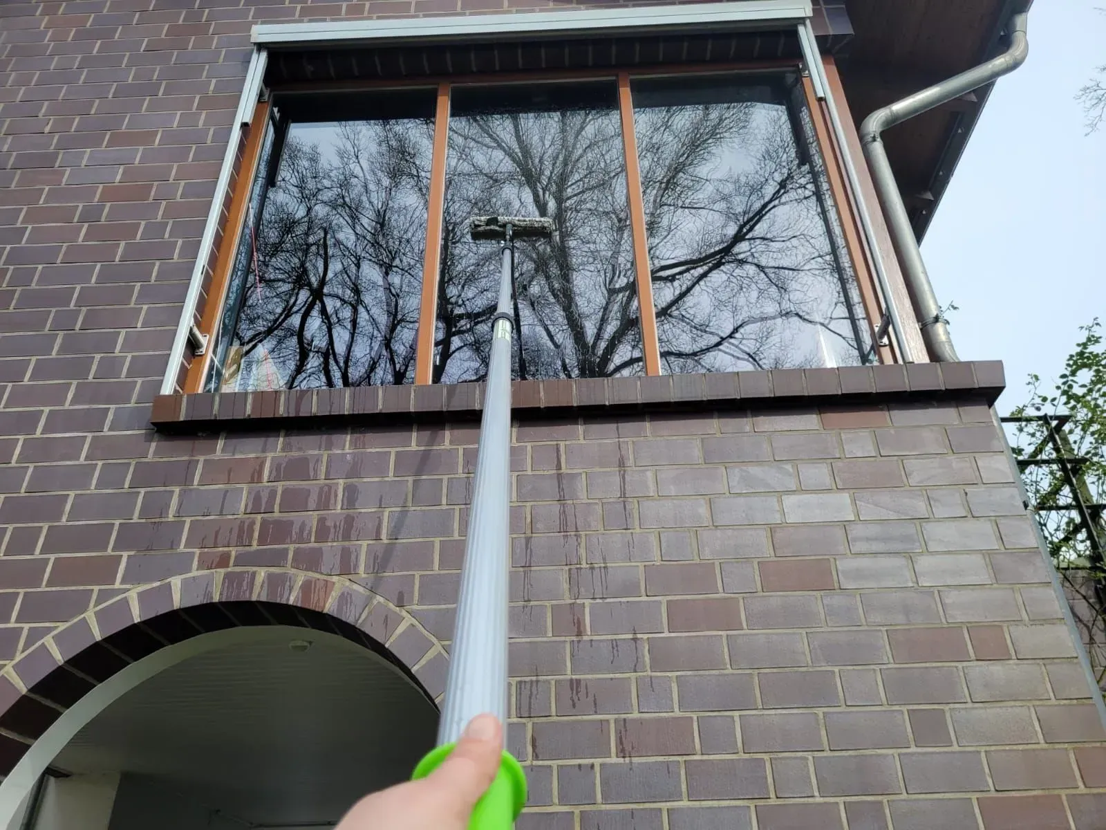 Grauer Pfahl, der bis zu einem Fenster eines Backsteingebäudes reicht, in dessen Scheibe sich Bäume spiegeln