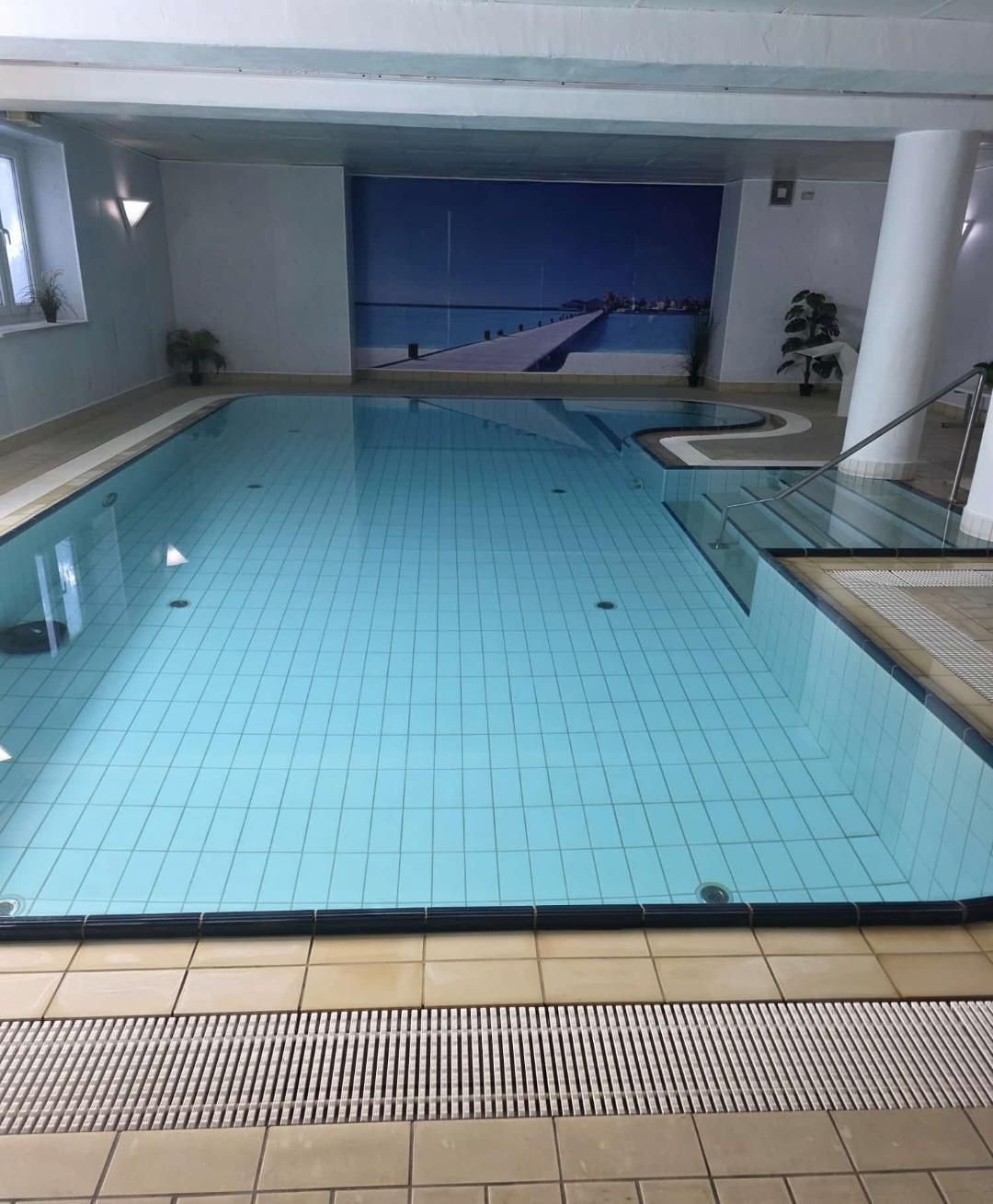 Hallenbad mit hellblauem Wasser, gefliestem Beckenrand und einer Wand mit einem Wandgemälde im Hintergrund