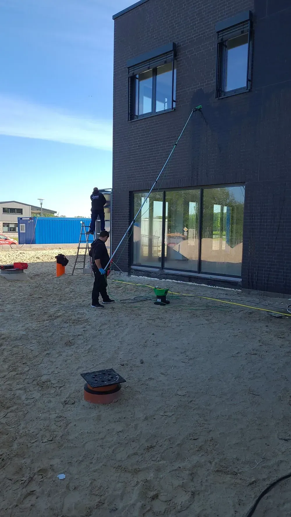 Eine Person arbeitet neben einem dunklen, zweistöckigen Haus im Bau. Rundherum stehen Gerüste und Werkzeuge auf dem Boden.