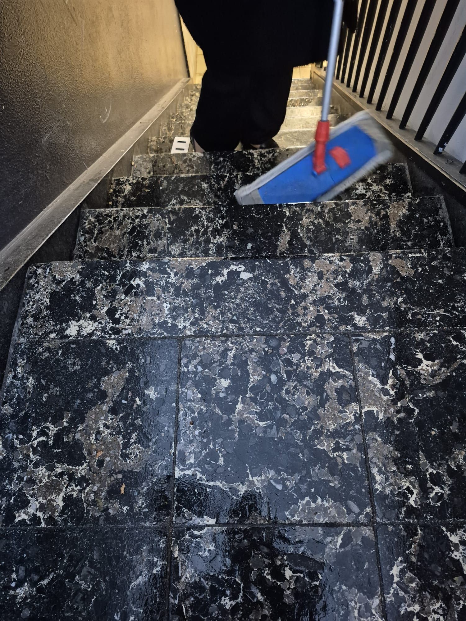 Person, die einen dunklen, gefliesten Treppenabsatz mit Kehrblech und Besen reinigt; auf dem Boden liegen verstreute Krümel
