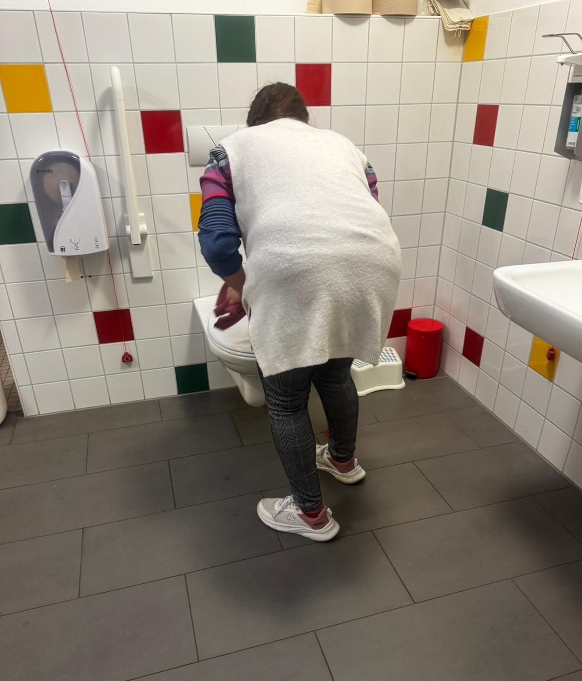 Person, die einem Kind an einem niedrigen Waschbecken in einer gefliesten Toilette mit bunten quadratischen Fliesen hilft