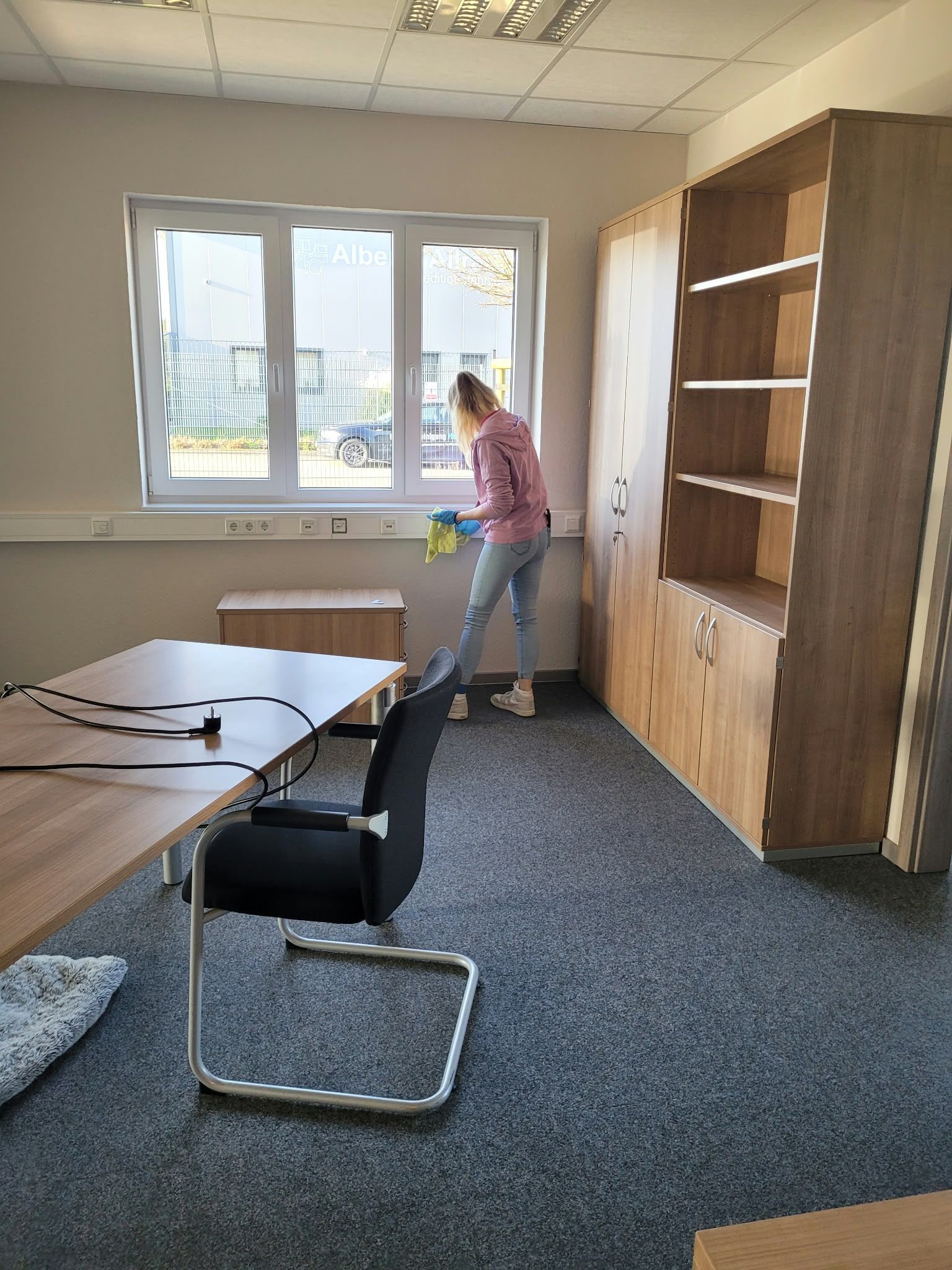 Büro mit Schreibtischen und Schränken; eine Person steht am Fenster und putzt oder ordnet Gegenstände.