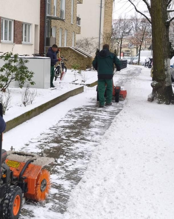 Arbeiter räumen mit orangefarbenen Schneefräsen einen Gehweg neben Wohnhäusern frei.